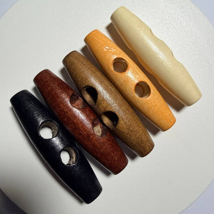 Four elongated wooden beads with circular cutouts, shown in black, reddish-brown, medium brown, and cream, arranged diagonally on a white textured surface.